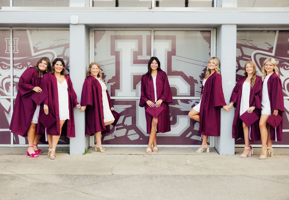 Graduates in maroon robes posing along a concrete wall for group photos.