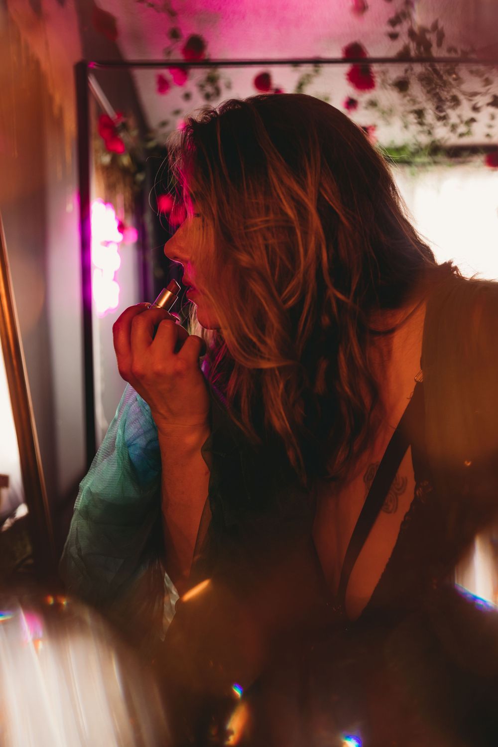 A silhouetted figure applies lipstick in moody lighting with pink reflections and prismatic light effects.