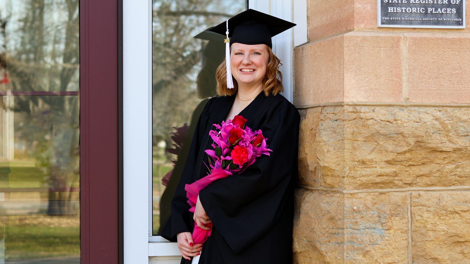 Graduate in cap and gown holding bright pink bouquet in front of brick building.