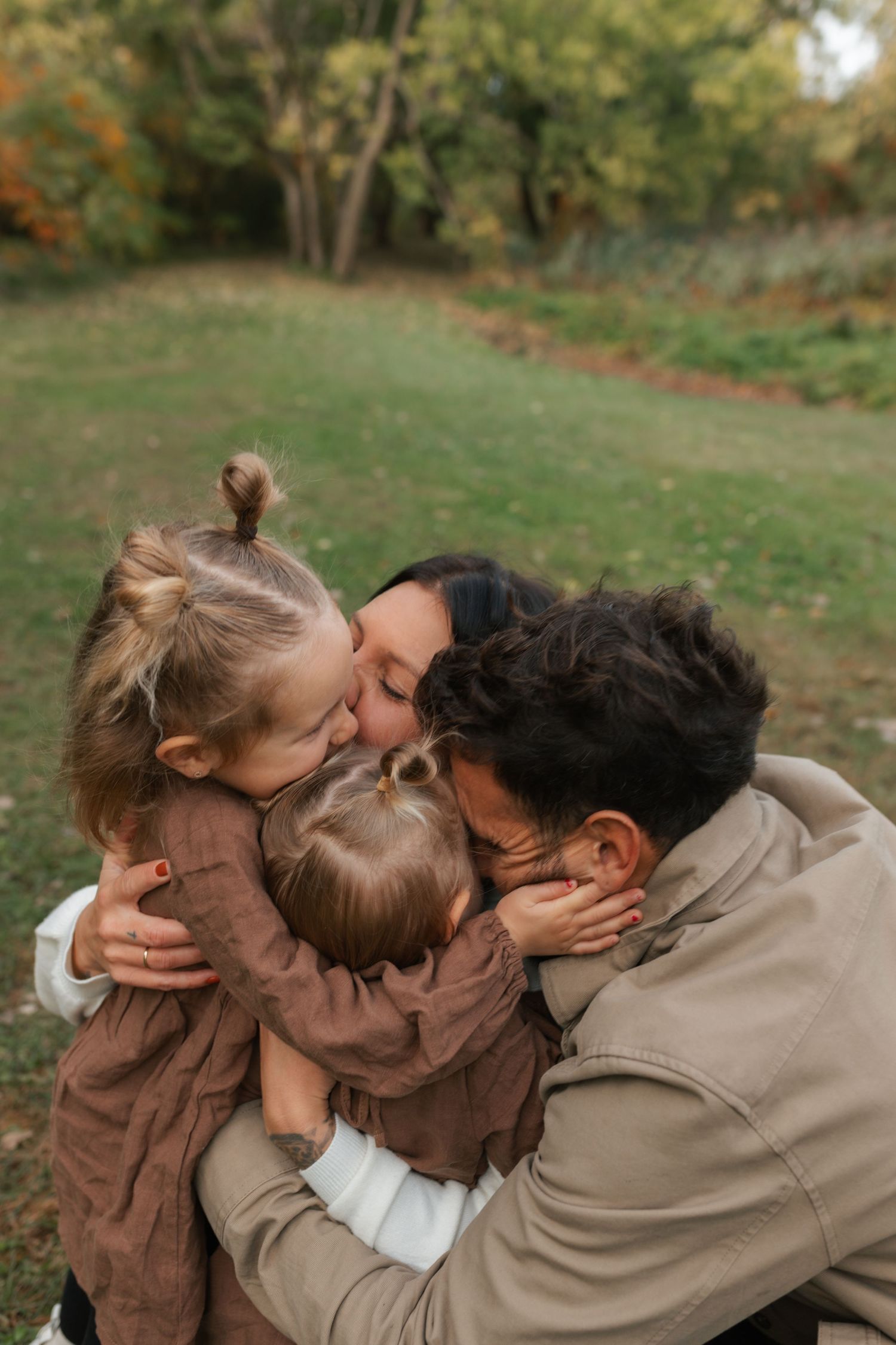 A joyful family shares a warm group hug and kisses in an autumn park, with two young daughters embracing their parents.