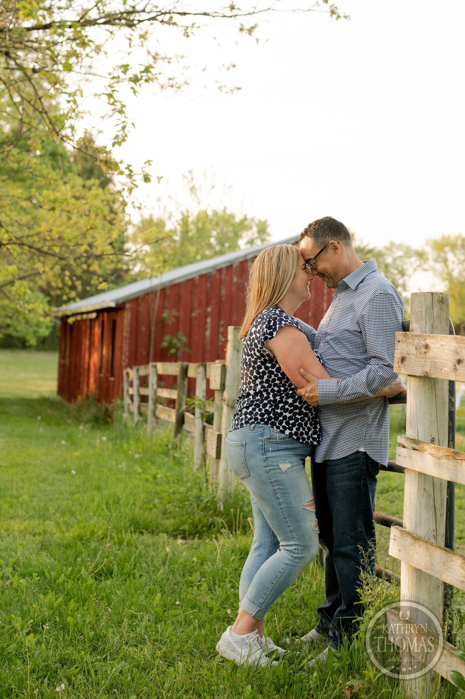 A couple shares an intimate moment by a wooden fence with a rustic red barn in the background.