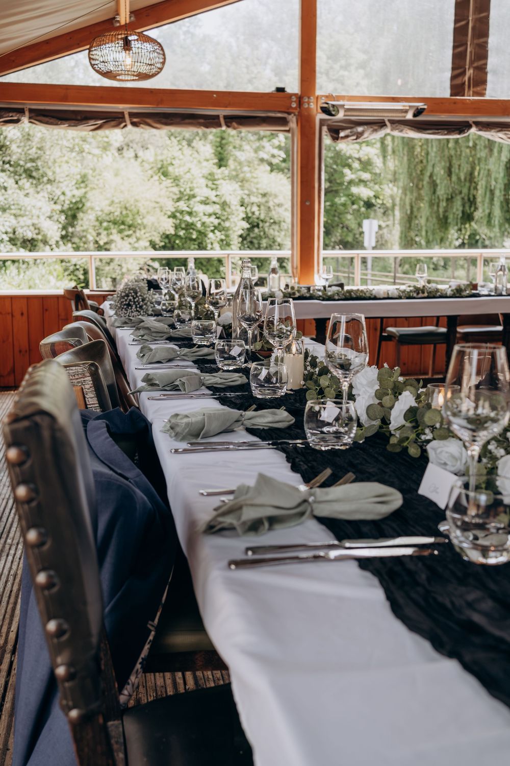 Elegant long banquet table set with black runner, green napkins, crystal glasses, and floral decor in a windowed venue.