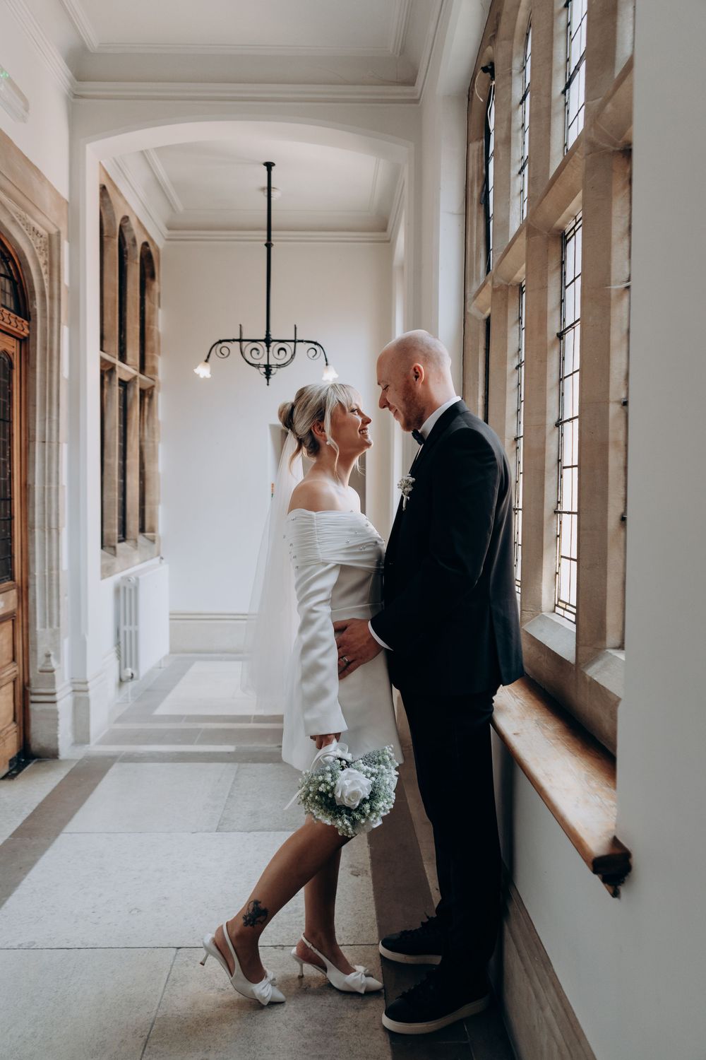 Bride and groom share a romantic moment in an elegant hallway, she holds a white bouquet wearing a short wedding dress.