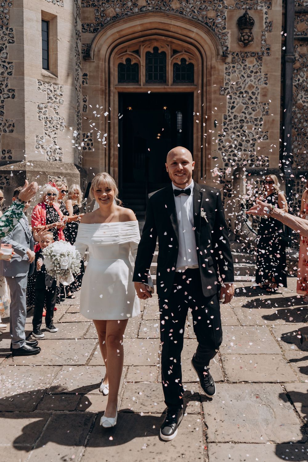 Bride in white mini dress and groom in black tuxedo exit church amid confetti shower from cheering guests.