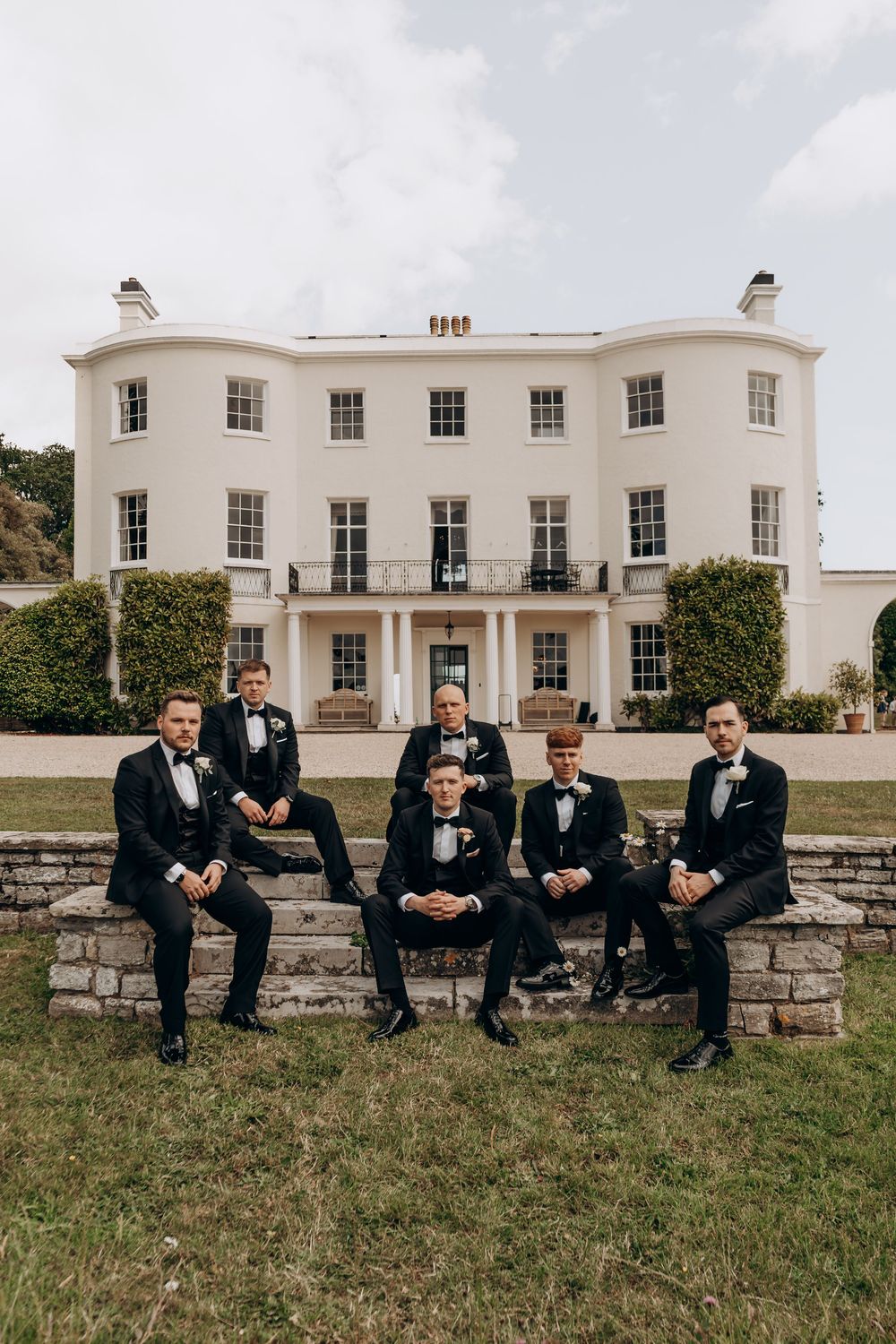 Six groomsmen in black tuxedos pose in front of an elegant white Georgian manor house on a wedding day.
