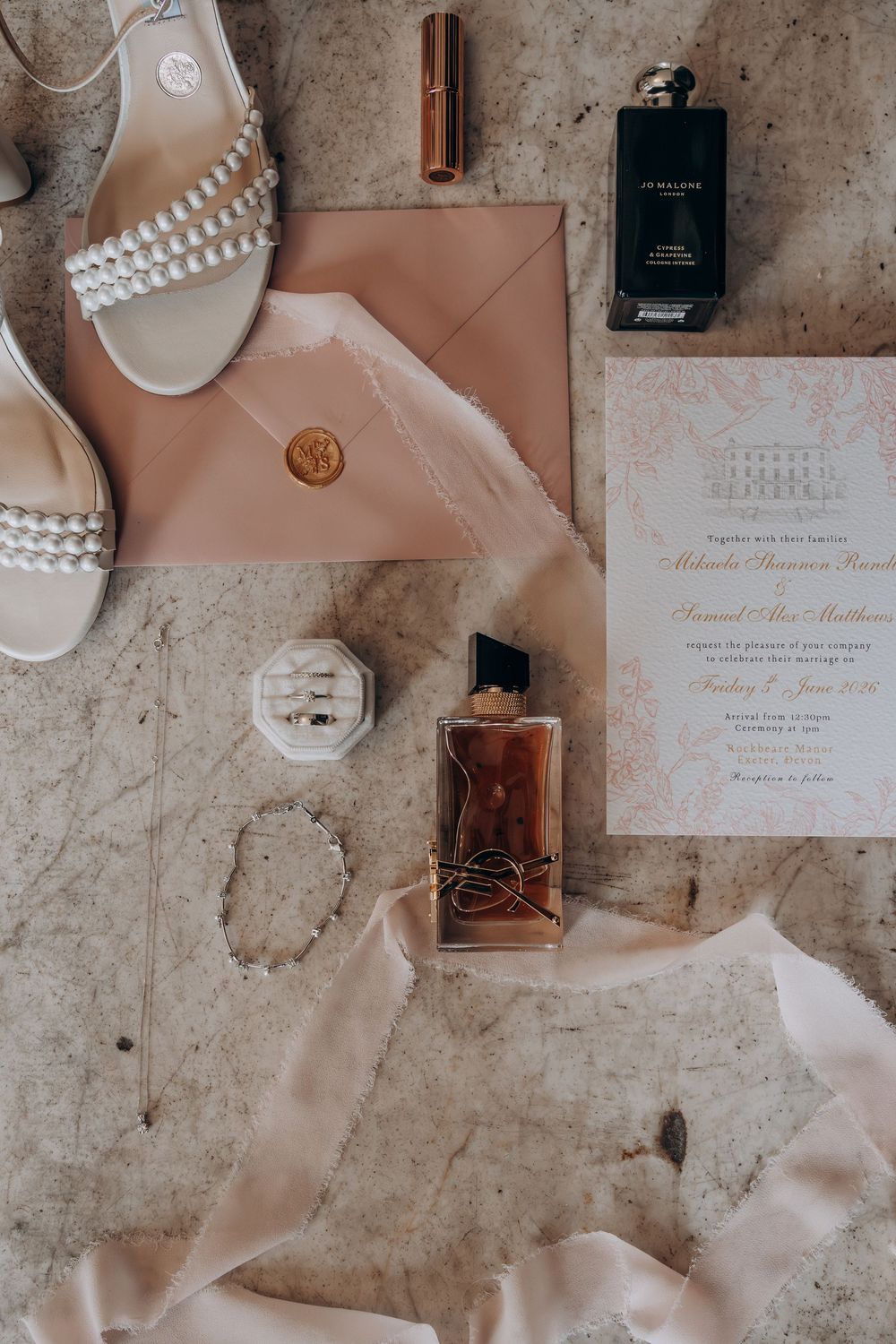 Elegant wedding flat lay with blush invitation, pearl heels, YSL perfume, Jo Malone fragrance, gold wax seal envelope, and rings.