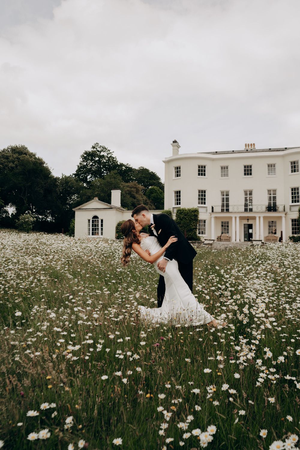 Bride and groom share a romantic dip kiss in a daisy field before a grand white Georgian manor house.
