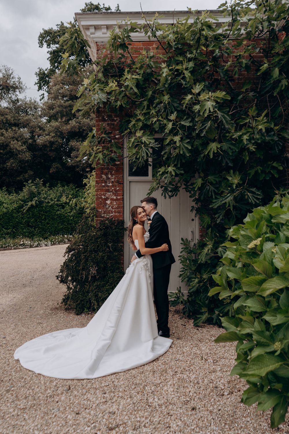 Bride and groom embracing in front of ivy-covered brick wall at elegant outdoor wedding venue.