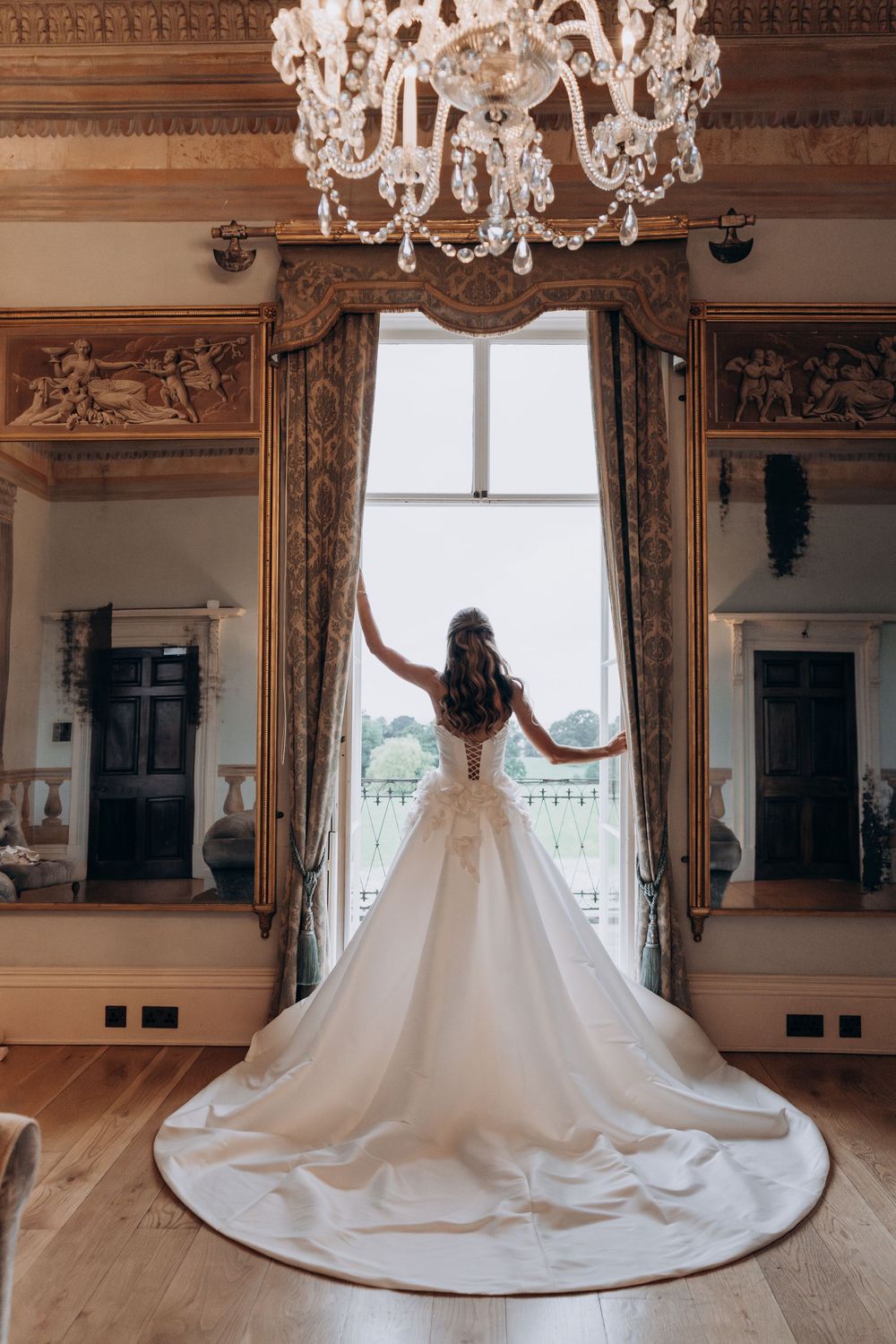 Bride in a stunning white ball gown with long train stands before an ornate window in an elegant chandelier-lit room.