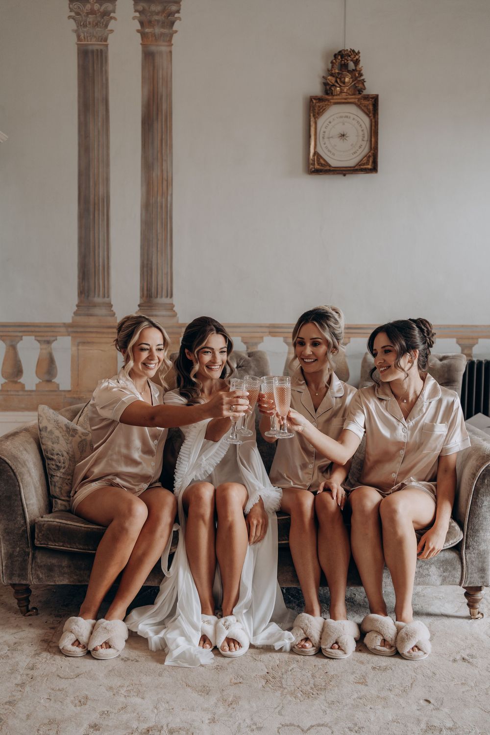 Four bridesmaids in satin pajamas and fluffy slippers toast champagne on a velvet sofa during wedding morning preparations.
