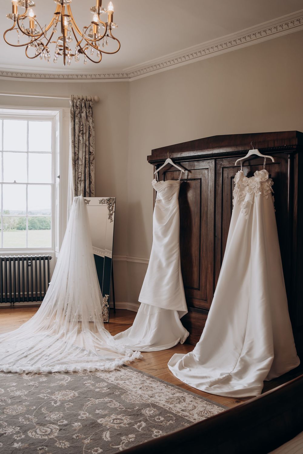 Three elegant white wedding dresses hanging in a classic room with chandelier and large window.