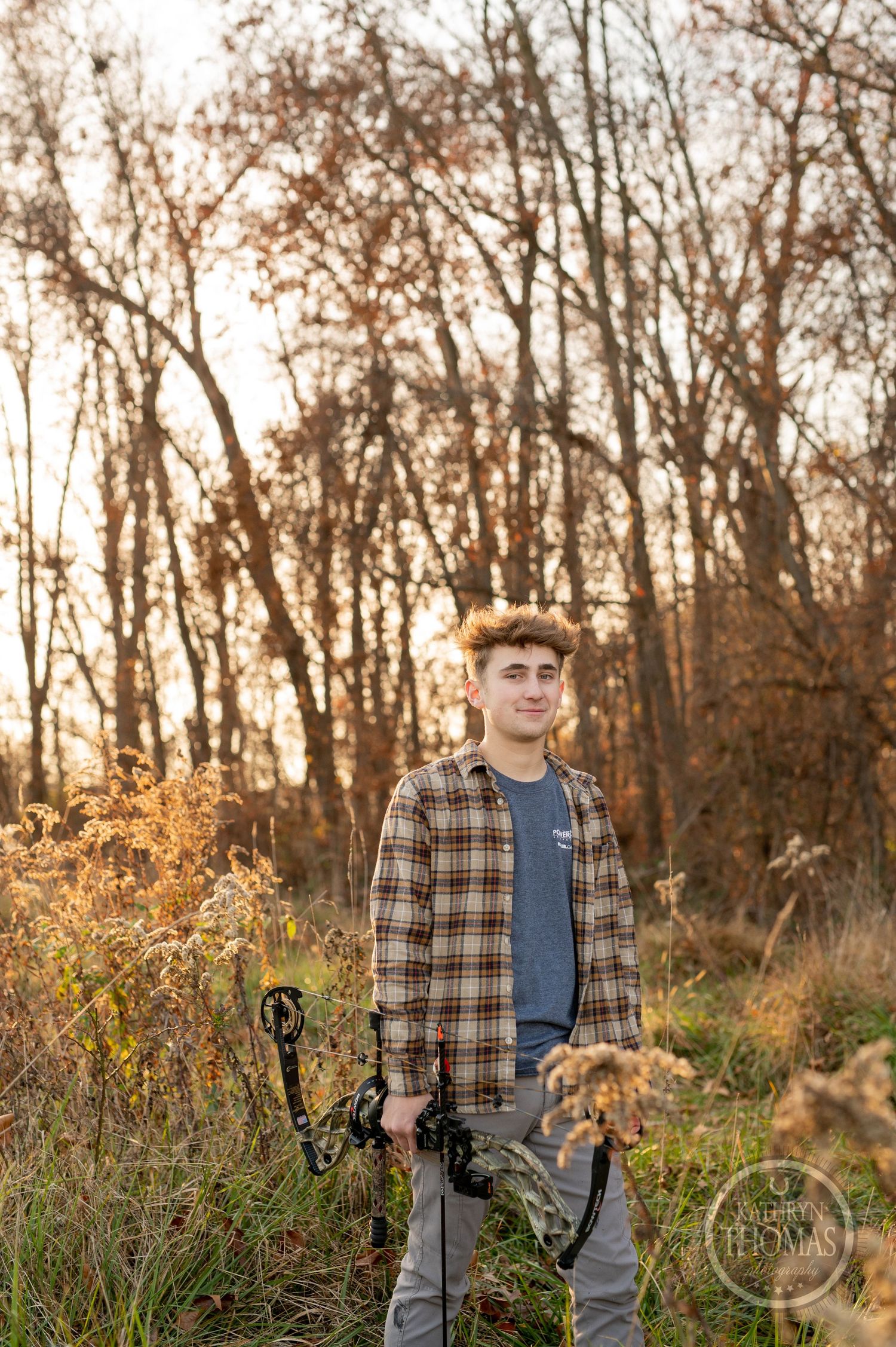 Archer in plaid shirt stands among autumn trees at sunset holding compound bow in grassy field.