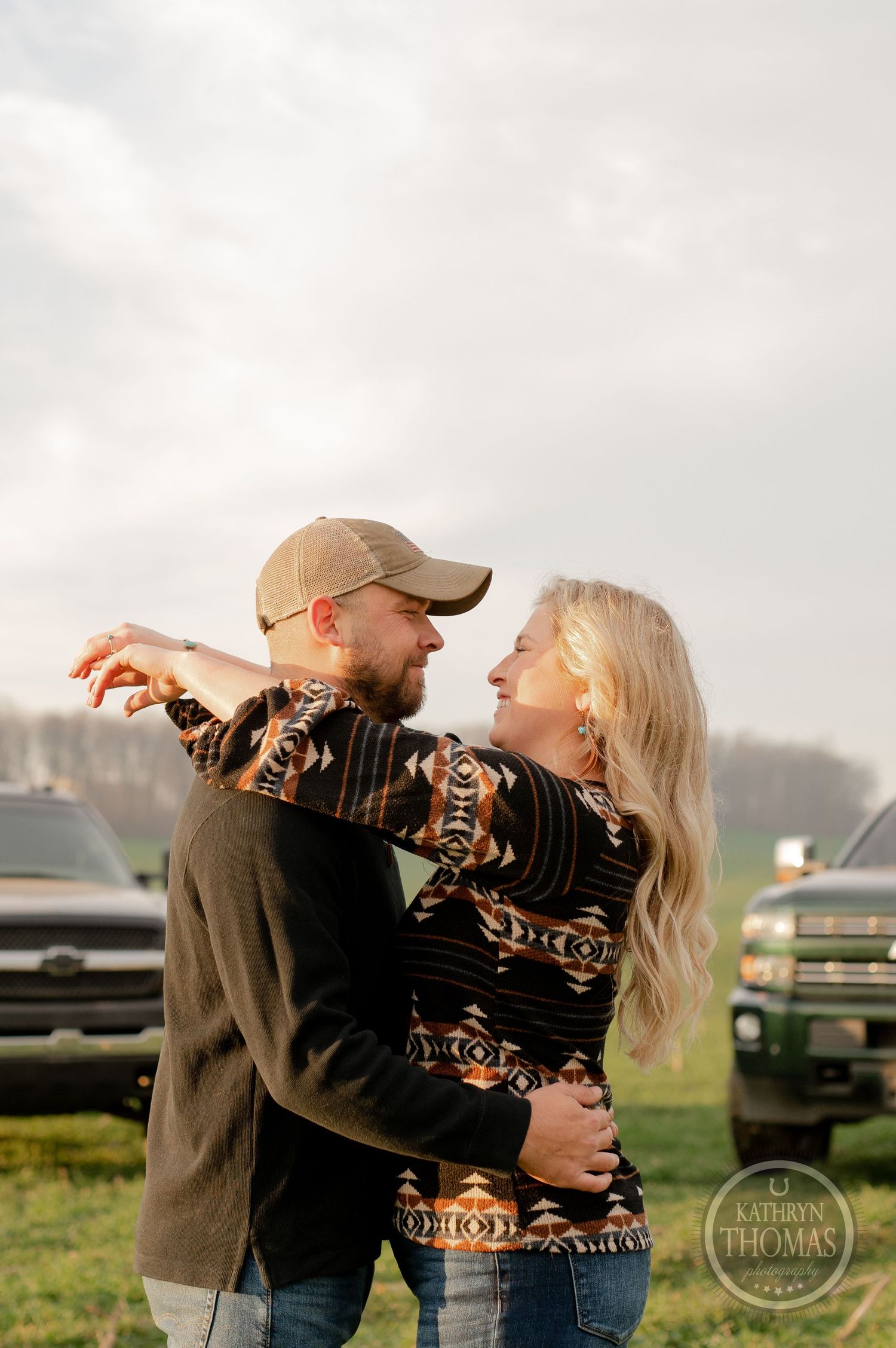 A couple in casual clothing embraces in front of pickup trucks in a grassy field.