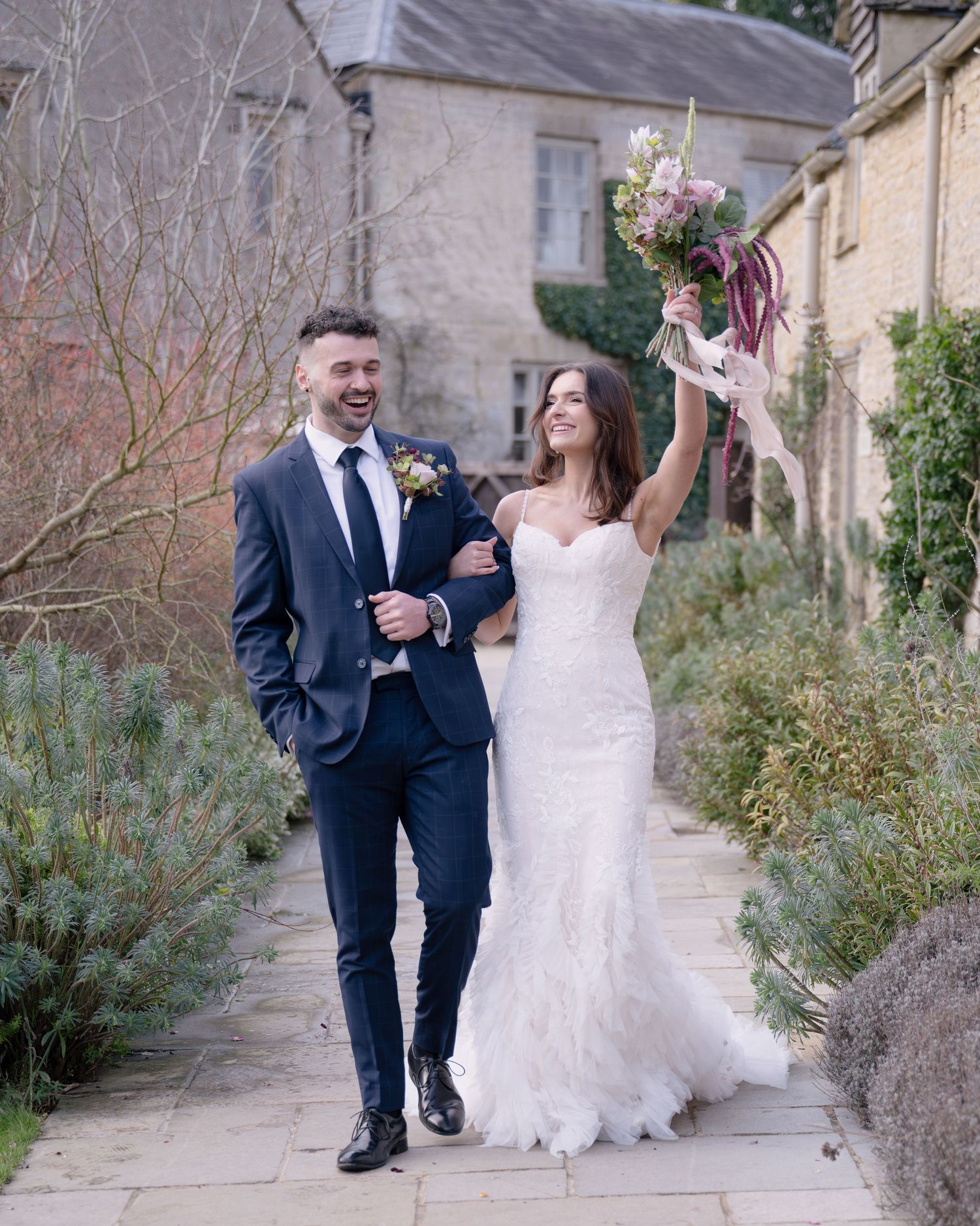 A joyful bride raises her bouquet while walking arm in arm with her groom outside a stone building.