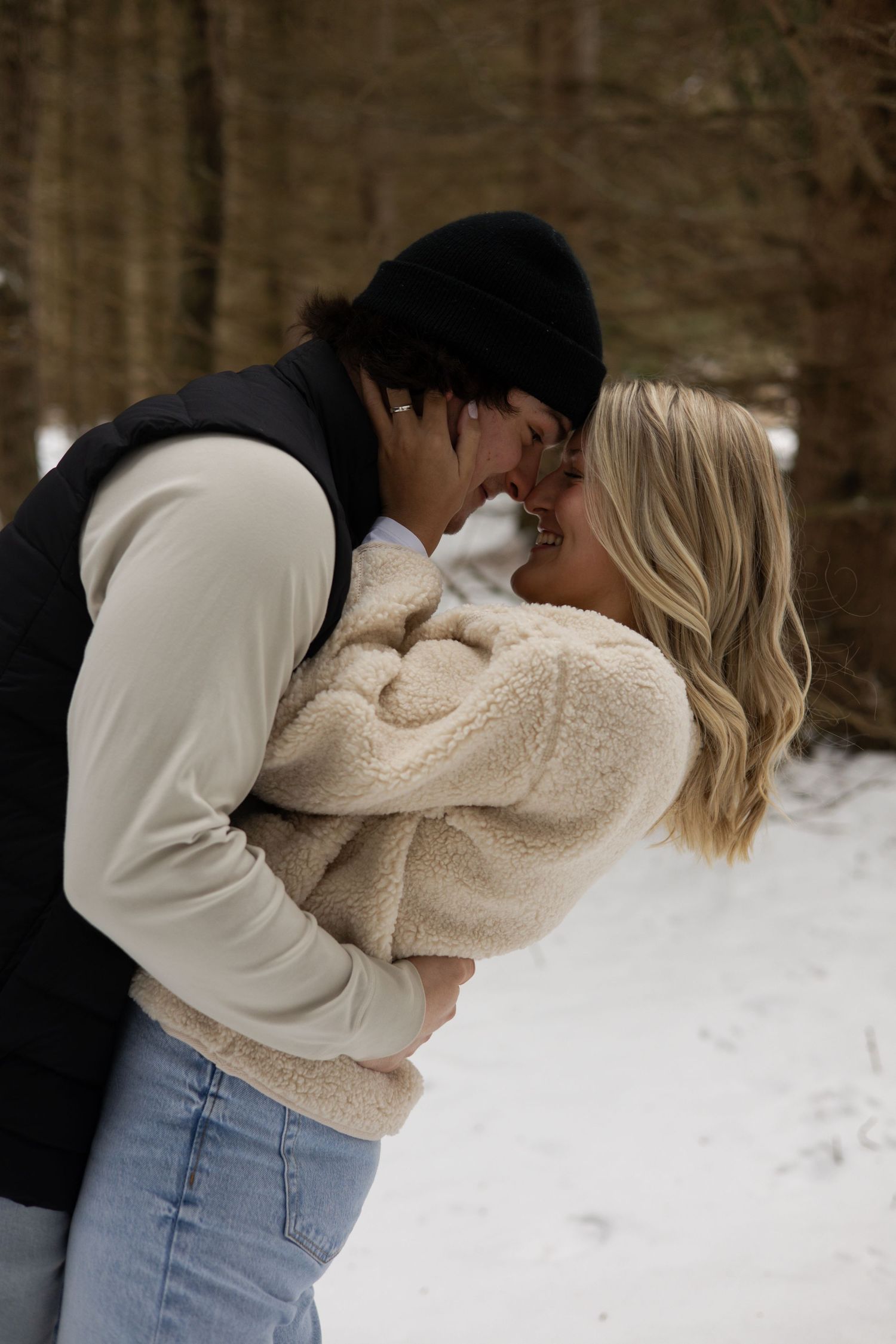 A couple shares an intimate moment outdoors in winter, with snow on the ground and trees in the background.