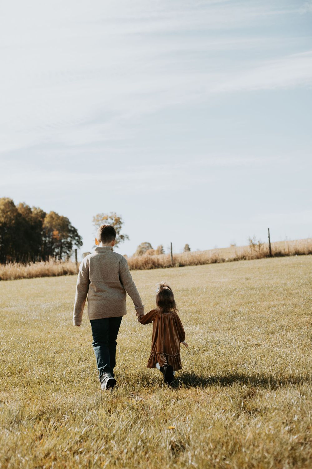 Two people walk together through a golden wheat field on a sunny autumn day with trees in the distance.