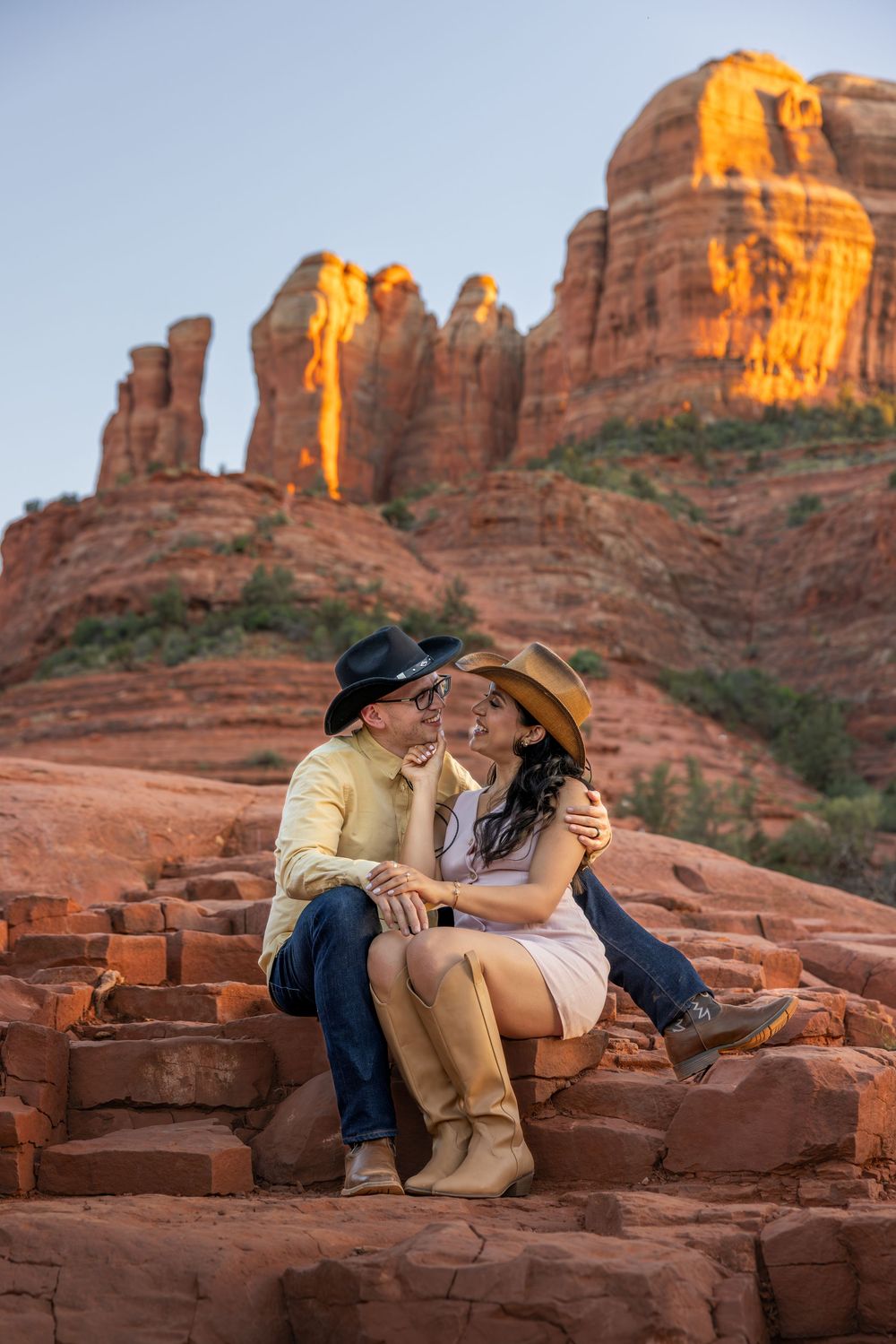 Couple in cowboy hats sitting close on red rocks in Sedona at sunset.
