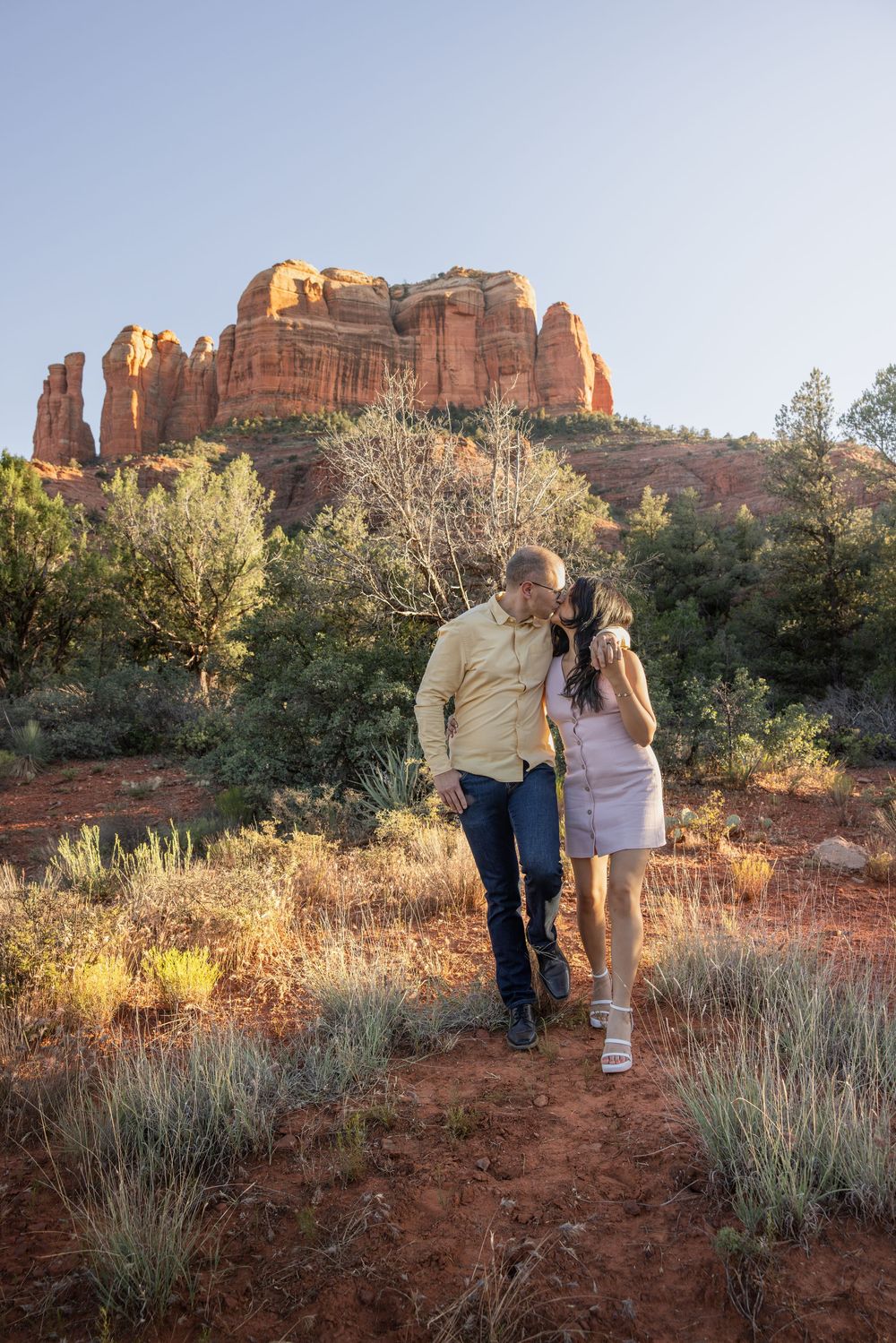 Couple shares a tender kiss while walking among Sedona red rock landscape in golden sunlight.