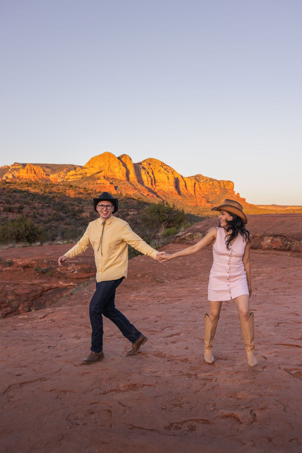 Playful couple holding hands and dancing on red rocks in Sedona at dusk.