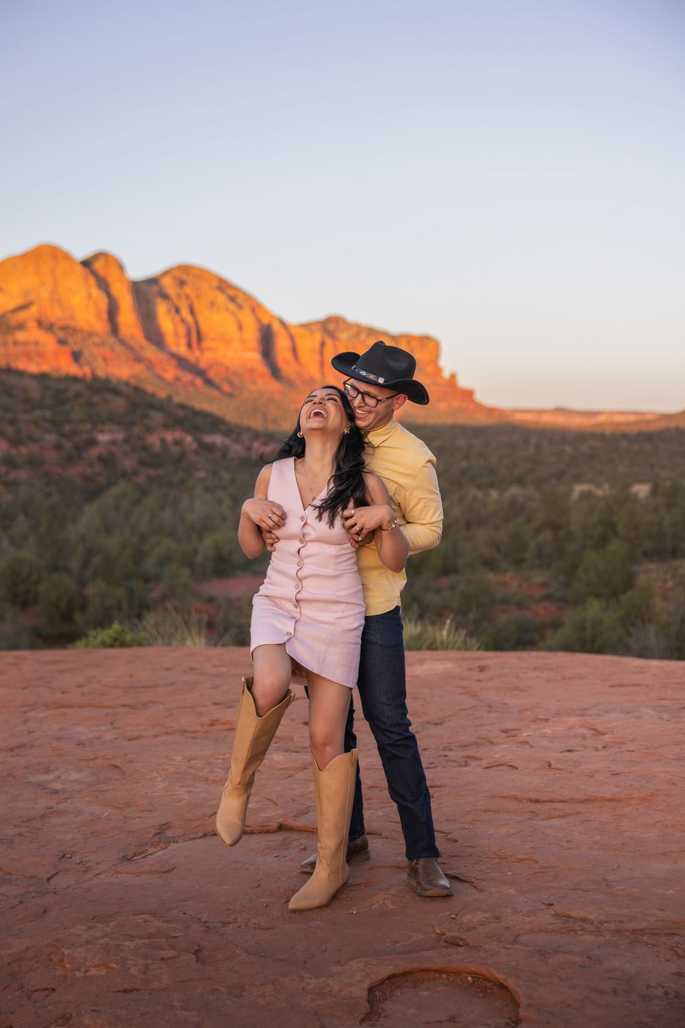 Couple laughing and embracing on red rocks with glowing Sedona mountains behind.