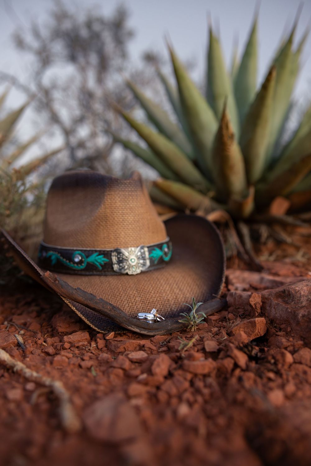 Brown cowboy hat with turquoise band resting on red desert ground near agave plant.