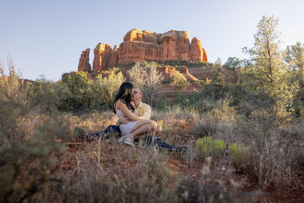 Couple sits closely together in desert brush with dramatic Sedona Cathedral Rock formation at sunset.
