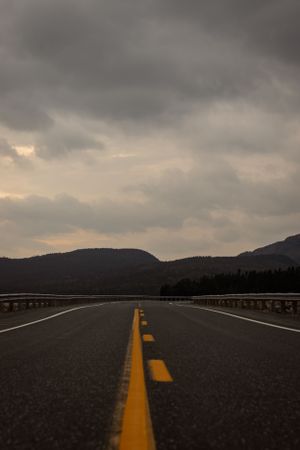 A winding mountain road with yellow dividing lines stretches into the distance under dramatic stormy clouds at dusk.