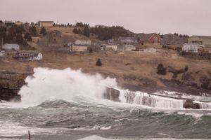 Large ocean waves crash against rocky coastline near seaside homes on a misty hillside in coastal town.