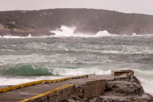 Large waves crash against a concrete pier during stormy weather on the Atlantic coast with forested hills in background.