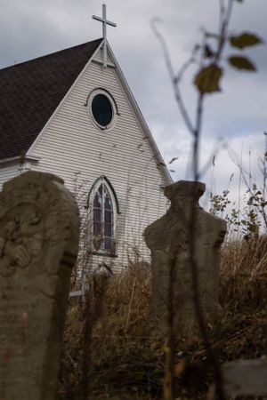Old weathered gravestones stand in front of a white rural church with Gothic windows and cross under moody skies.