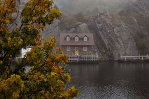 A rustic cottage with dormers sits on a misty lake surrounded by autumn foliage and rocky cliffs.