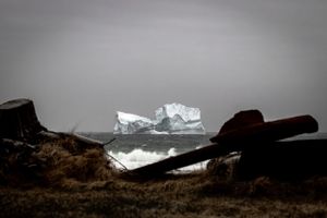 A large iceberg looms in the distance behind dark rocky outcrops on a moody, overcast coastline.