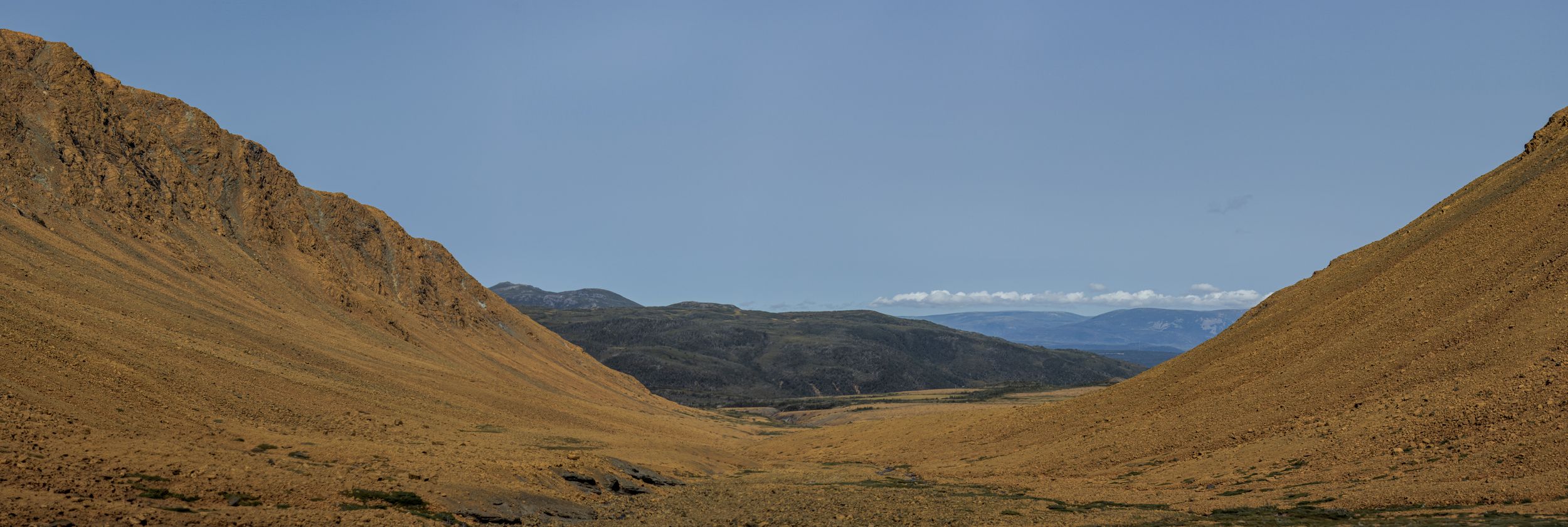 A panoramic mountain valley surrounded by brown rocky slopes and distant peaks under a clear sky.