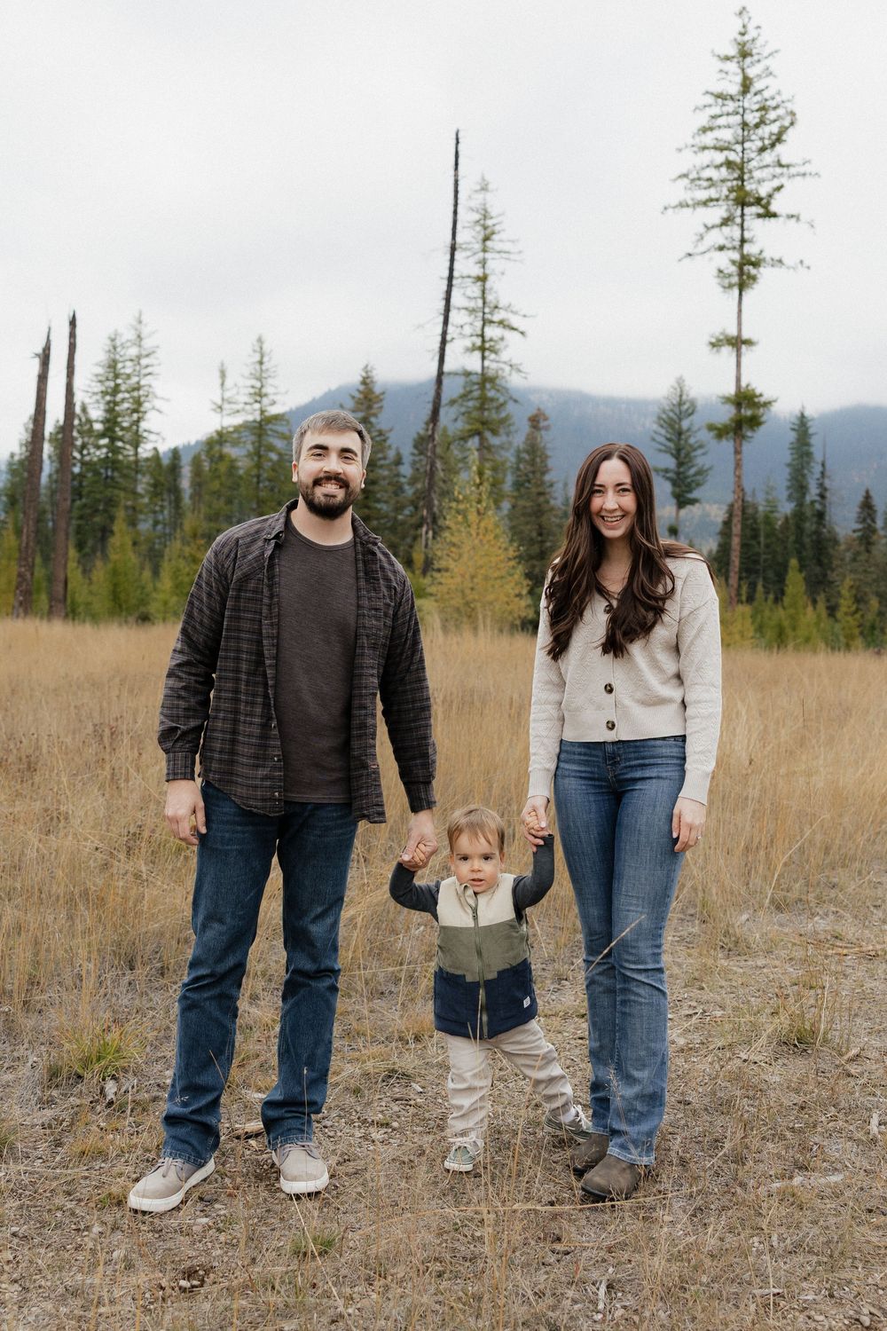 A young family with a toddler poses in a mountain meadow surrounded by pine trees and fall foliage.