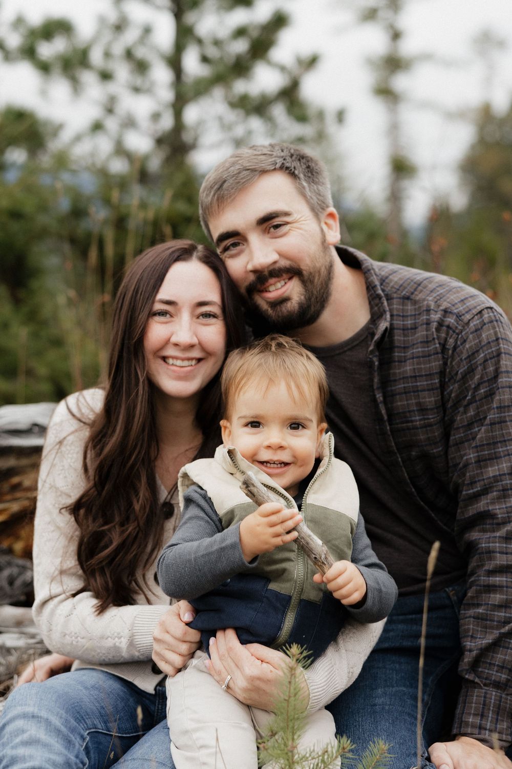 Smiling young family with a toddler posing together outdoors in a forest setting.