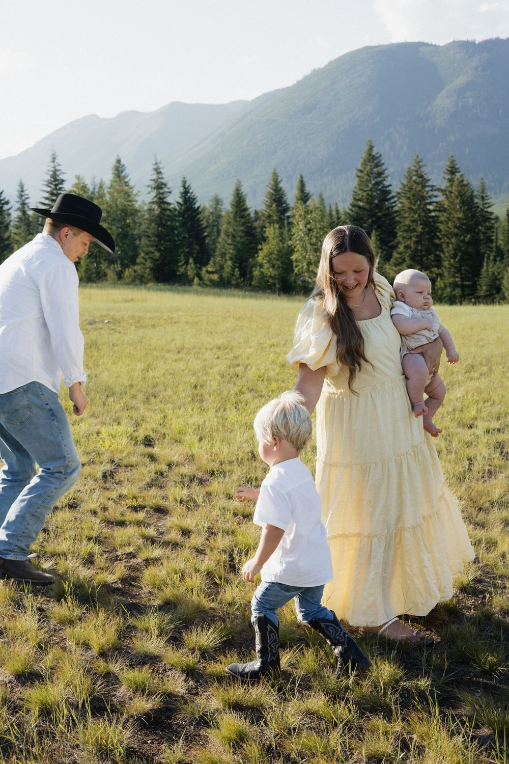 A family enjoys a sunny afternoon in a mountain meadow with evergreen trees and rolling hills in the background.