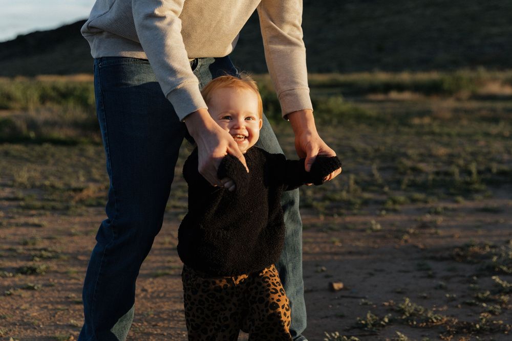 Adult helping a smiling toddler take first steps outdoors in golden hour sunlight on sandy ground.