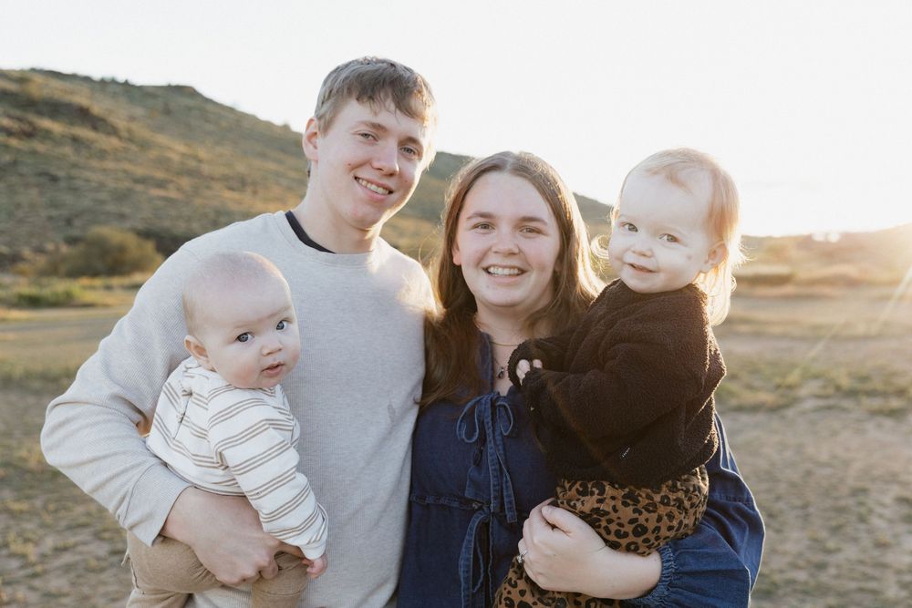 Young family smiling outdoors at golden hour, parents holding two babies in a scenic field with hills.