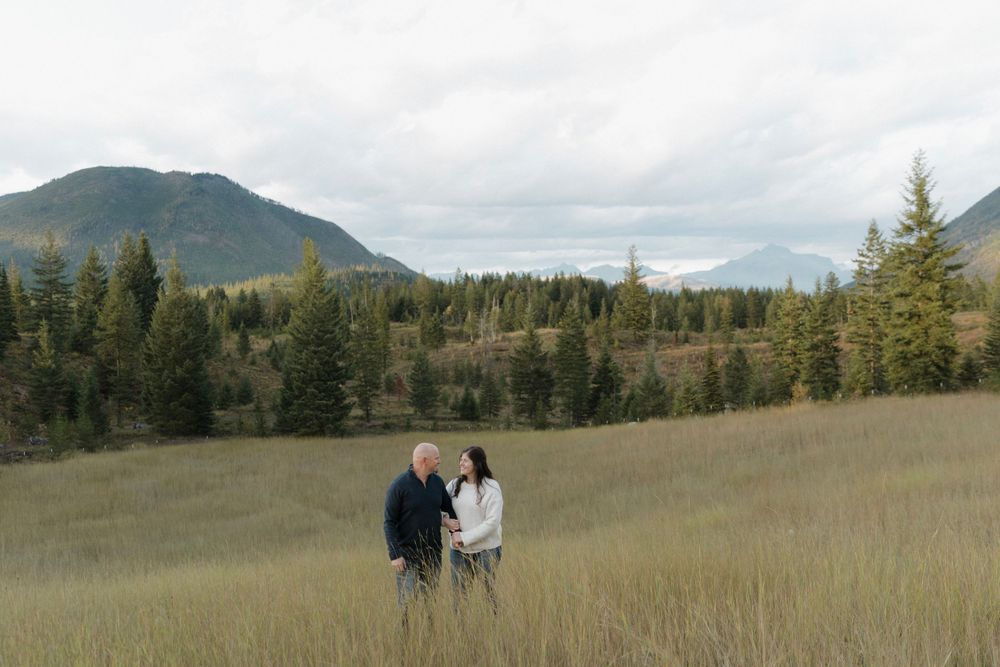 Couple stands in golden meadow with mountain views and evergreen forest.
