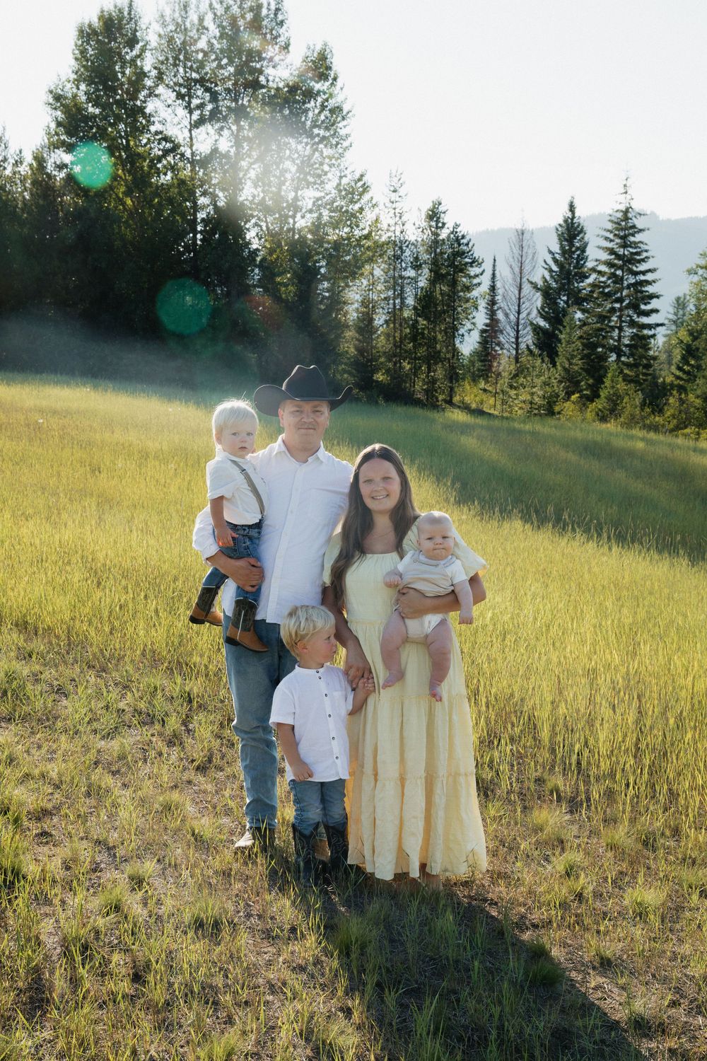 A family stands in a sunlit meadow with evergreen trees and mountains in the background on a bright summer day.