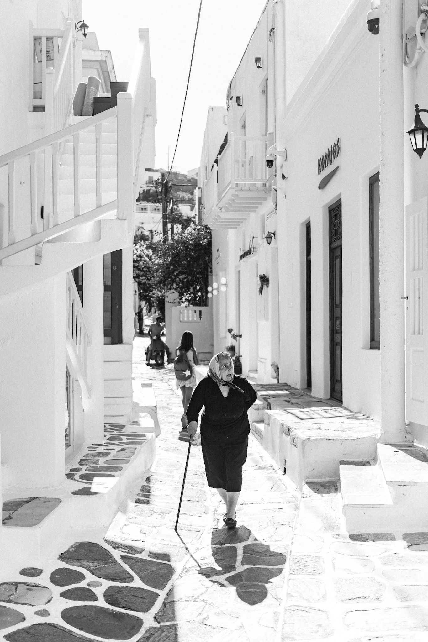 Person walking with a cane through a narrow whitewashed Mediterranean street with cobblestones.
