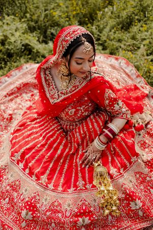 Traditional Indian bride in ornate red and gold lehenga sits elegantly with henna-adorned hands amid lush greenery.