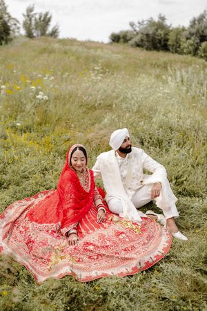 A couple in traditional Sikh wedding attire sits together on a grassy hillside with wildflowers in the background.