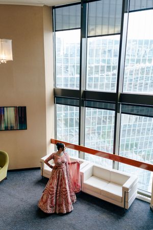 Inside a modern high-rise building, a figure in an ornate red and pink traditional lehenga stands near tall windows and white sofa.