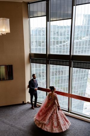 A couple in elegant South Asian wedding attire twirls and poses in front of floor-to-ceiling windows in a modern room.