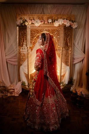 A bride in a luxurious red and gold embroidered Indian lehenga stands in front of an ornate golden backdrop with white flowers.