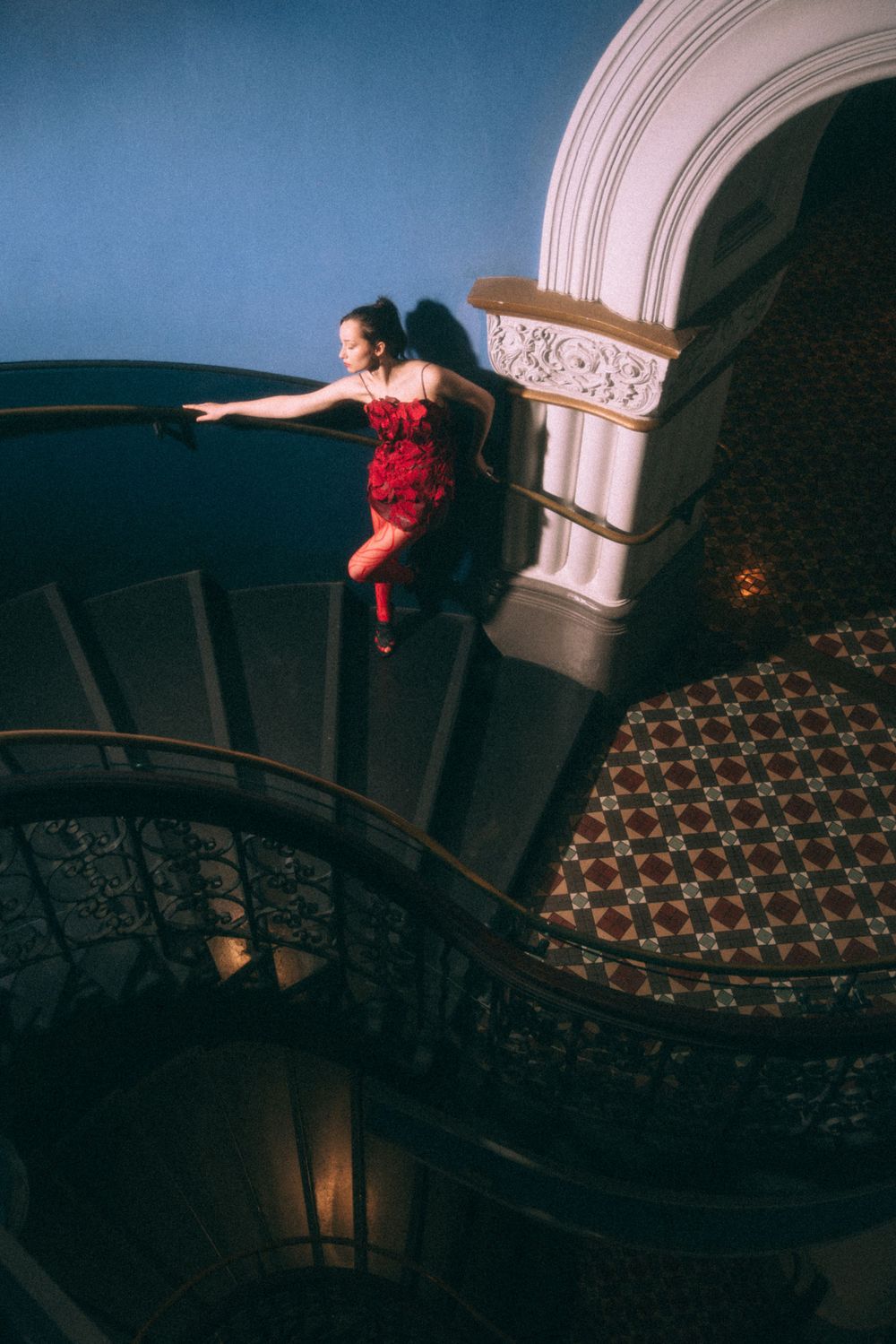 A dancer in a red dress performs a dramatic pose on an ornate architectural balcony with dramatic lighting and patterns.
