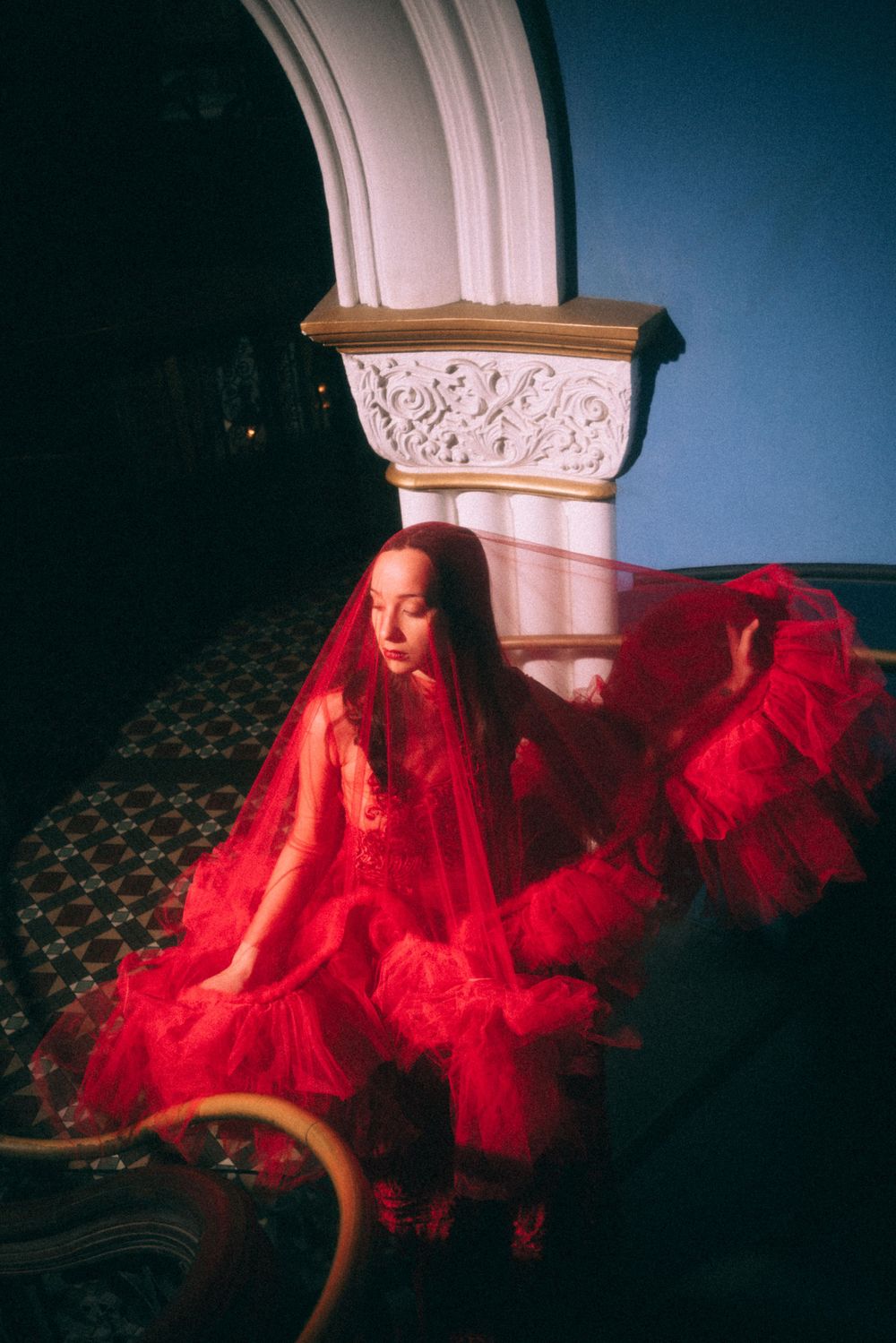 A figure in a flowing red dress and veil sits dramatically beside an ornate white column with carved details.