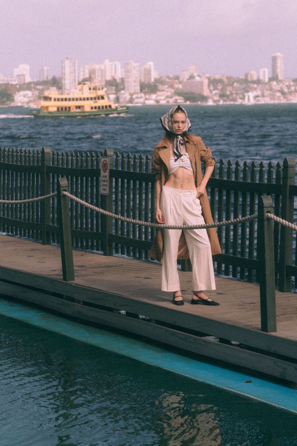 Figure in vintage-style outfit stands on harbor boardwalk with ferry boat and city skyline in background.