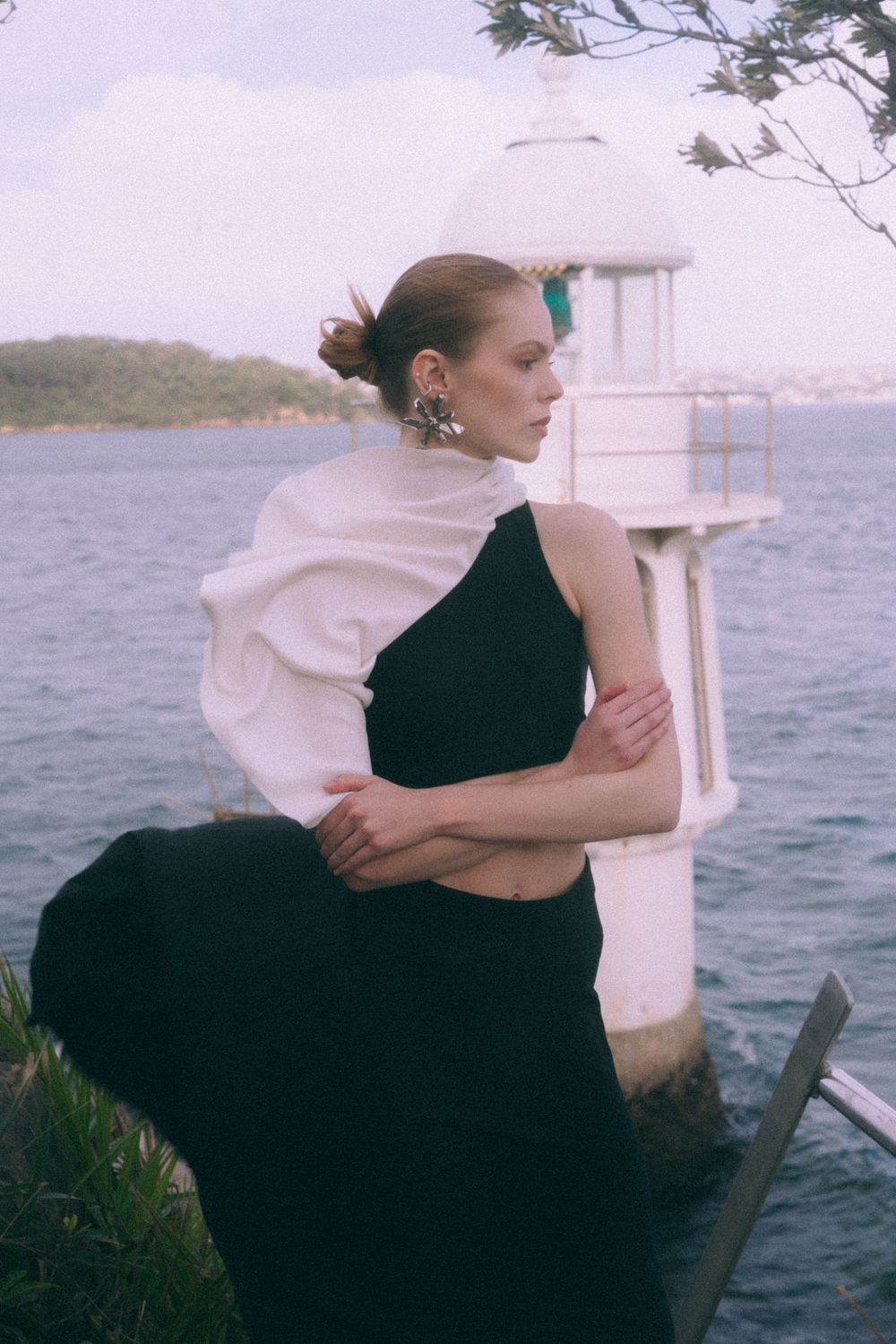 Elegant figure in black dress poses against coastal lighthouse and ocean backdrop in romantic vintage-style photo.